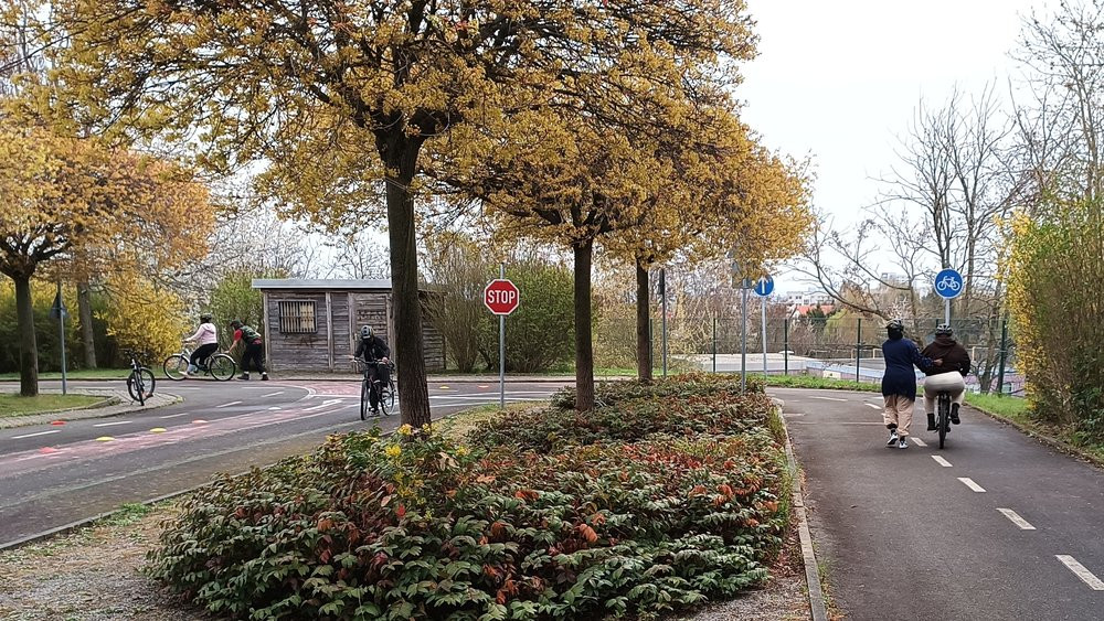 A photo of a training facility for traffic. There are three persons on bikes. Some of them are going alone, some with support by other persons. Between the little roads there are trees in yellow bloom.