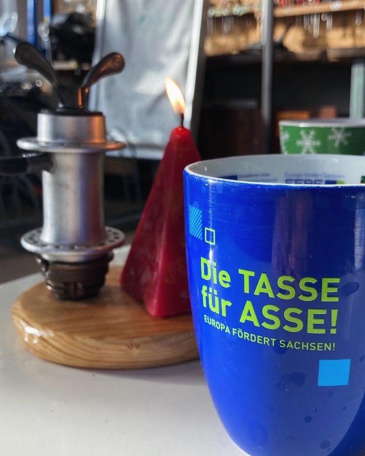 Eine blaue Tasse: Die Tasse für Asse - Europa fördert Sachsen. Dahinter eine brennende Kerze.