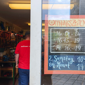 Eingang zum Rad i.O., links sieht man durch die offene Tür die Werkzeugwand und einen Menschen mit rotem Shirt. Rechts eine Tafel mit Öffnungszeiten.